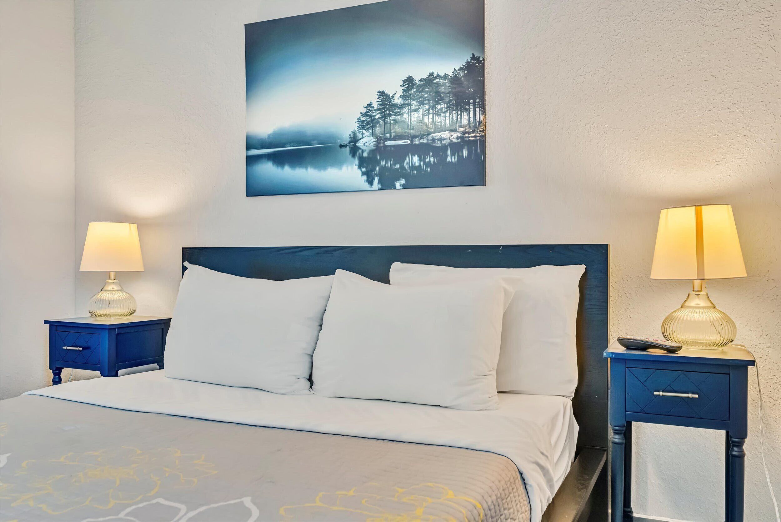 A neatly made bed with white bedding, two bedside tables with lamps, and a framed picture of a forest on the wall.