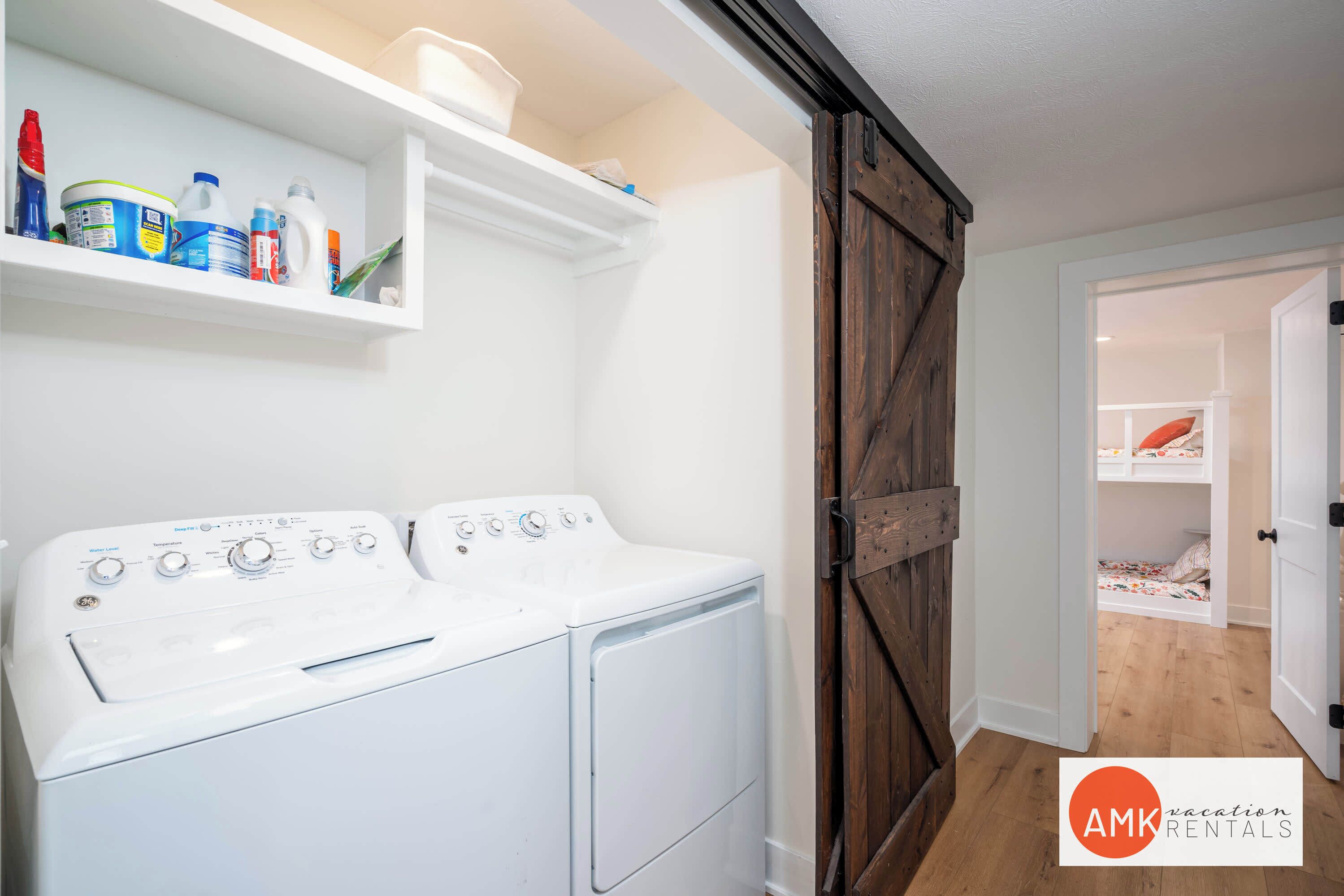 Un espace buanderie équipé d’un lave-linge et d’un sèche-linge à chargement frontal, d’une étagère contenant des produits de nettoyage et d’une porte en bois donnant sur une chambre avec un lit superposé.