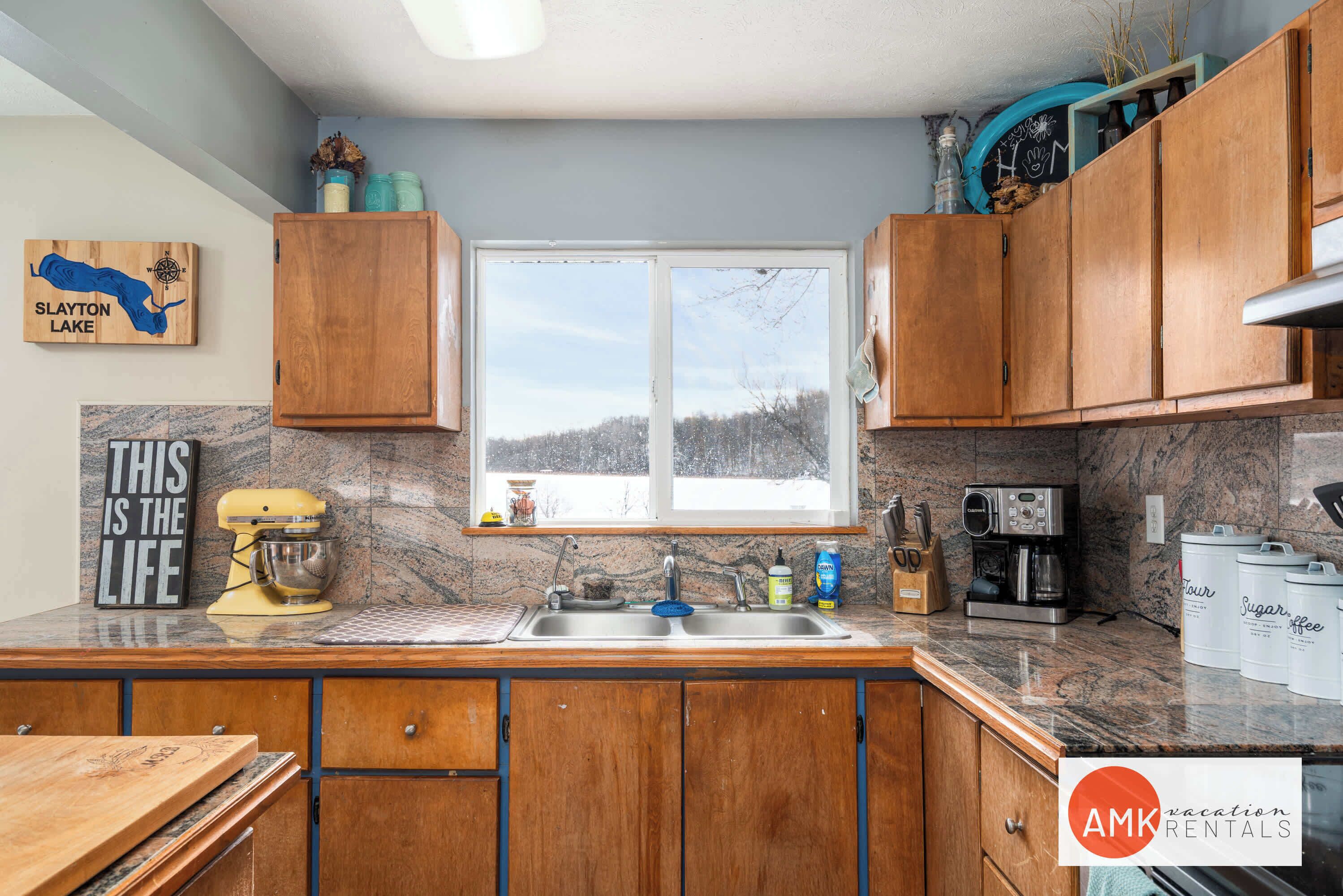 A kitchen with wooden cabinets, a granite countertop, a mixer, a coffee maker, and a window with a view of trees.
