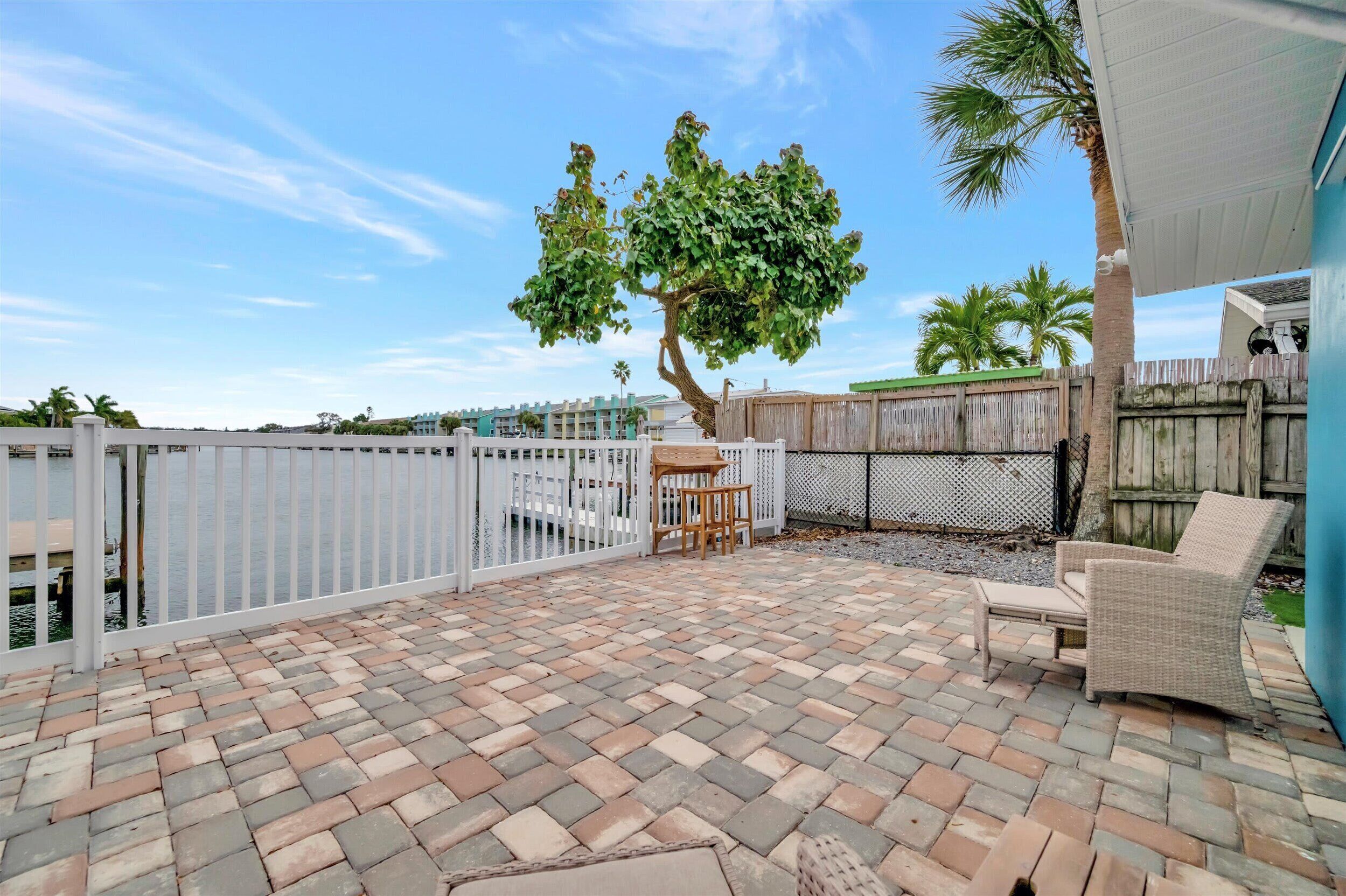A patio with a brick floor, a white fence, a tree, and outdoor furniture.