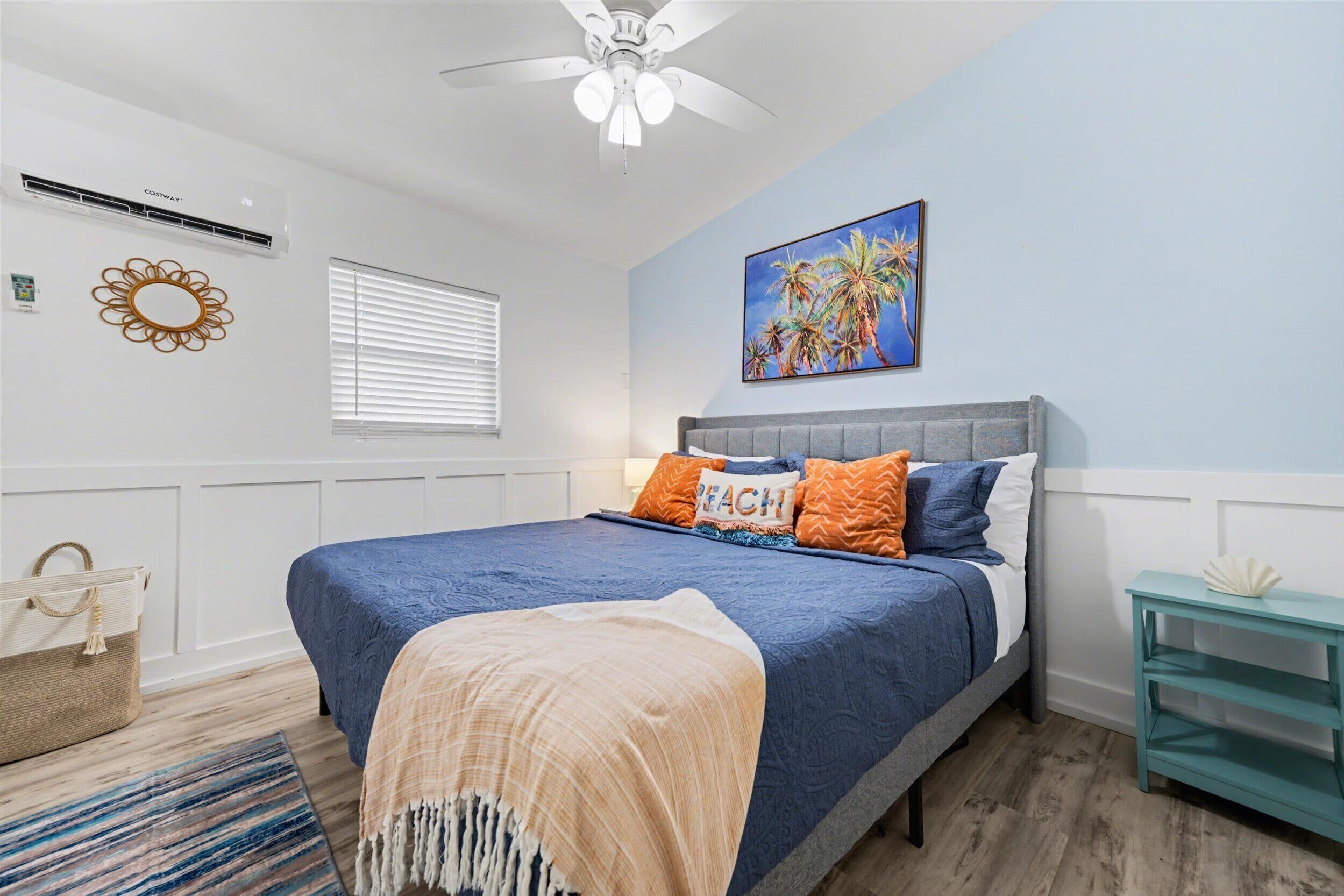 Un dormitorio con cama, ventilador de techo, aire acondicionado instalado en la pared, ventana con persianas, un cuadro y una pequeña mesa con un objeto decorativo.