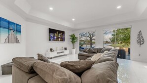 A modern living room with a sectional sofa, a white entertainment unit, and a large window offering a view of a body of water and greenery. - Waterview Villa Sleeps 10 BBQ Kayaks Games (Miami)