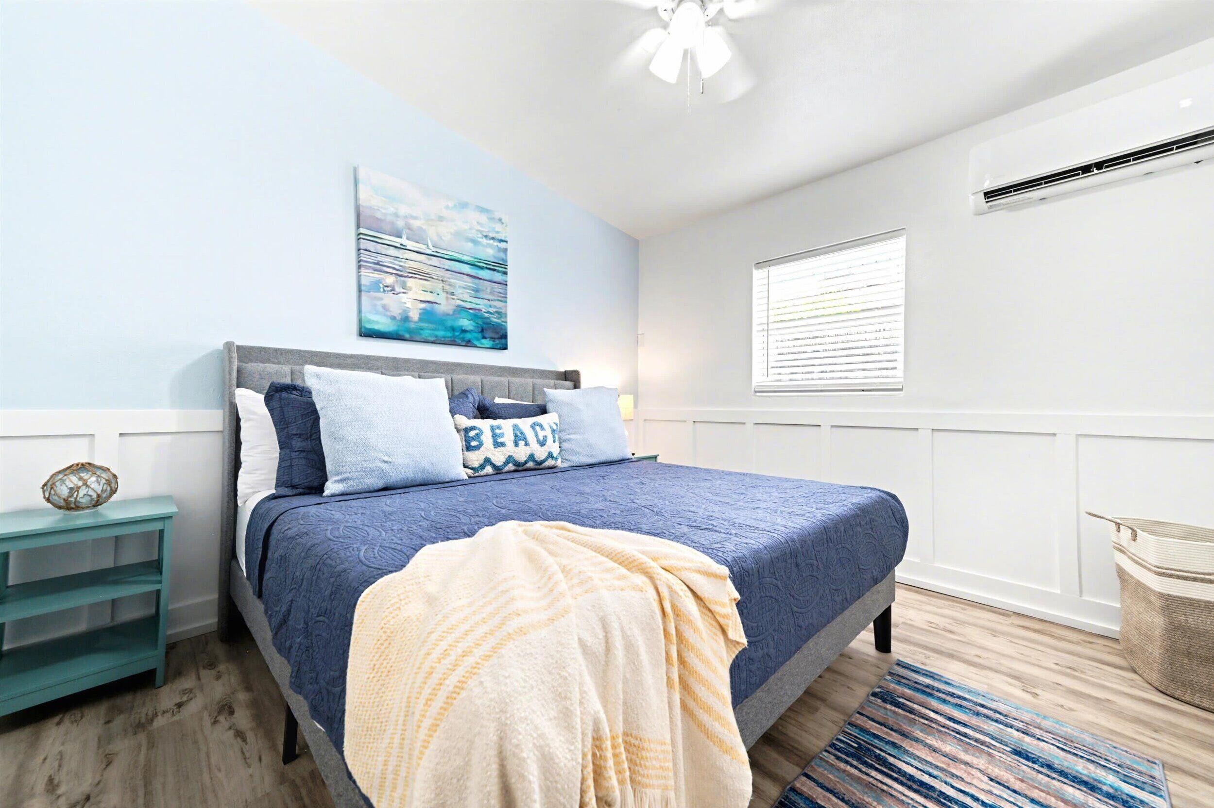 A bedroom with a bed, a nightstand, a wall-mounted air conditioner, a window with blinds, and a ceiling fan.