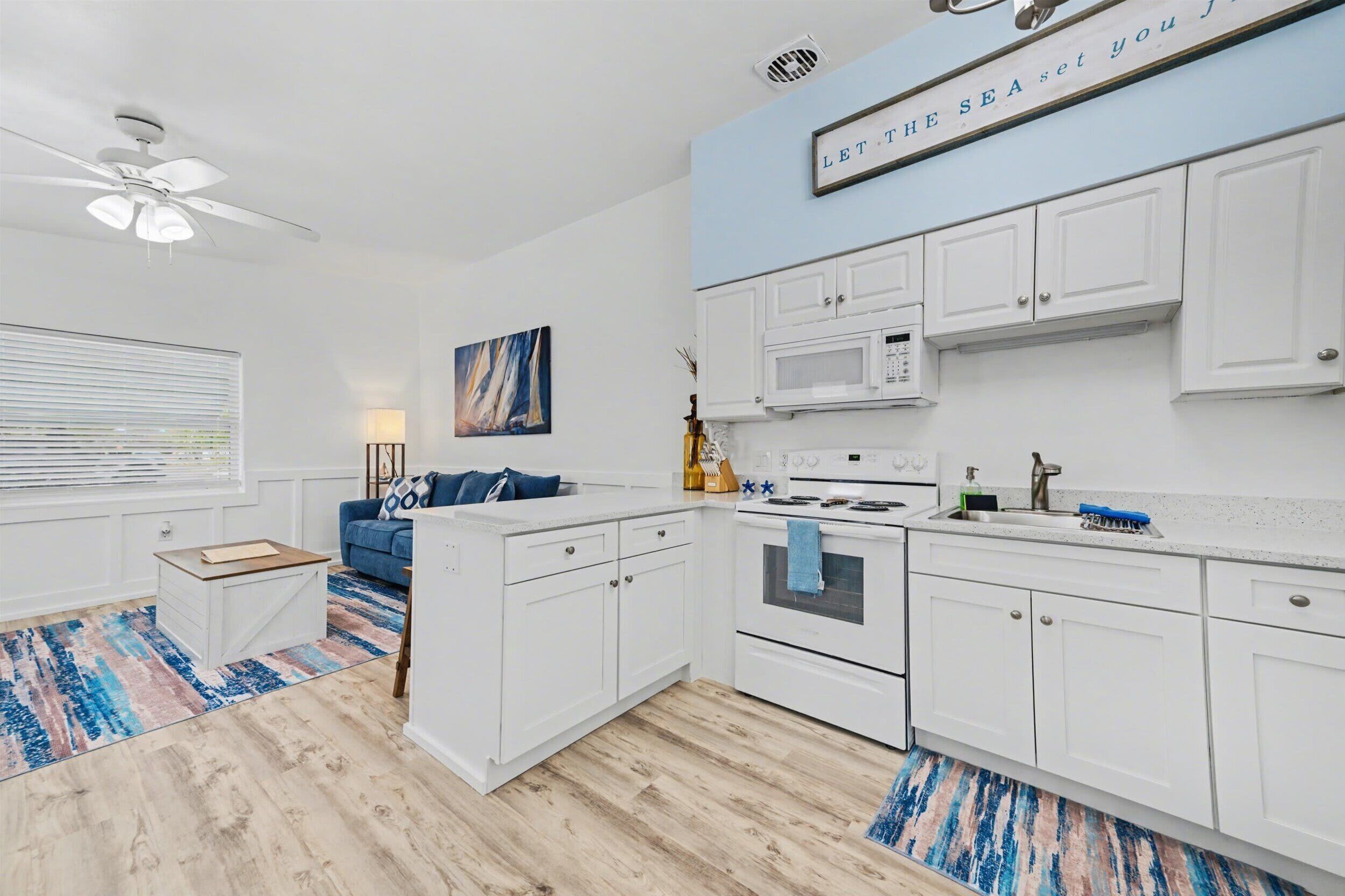 A modern kitchen with white cabinets, a microwave, and a stove. There's a blue painting on the wall and a ceiling fan.