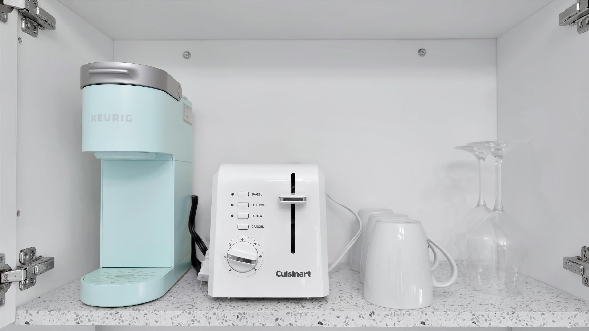 Eine Küchenzeile mit einer Keurig-Kaffeemaschine, einem Cuisinart-Toaster und zwei weißen Tassen auf einer fleckigen Arbeitsplatte.