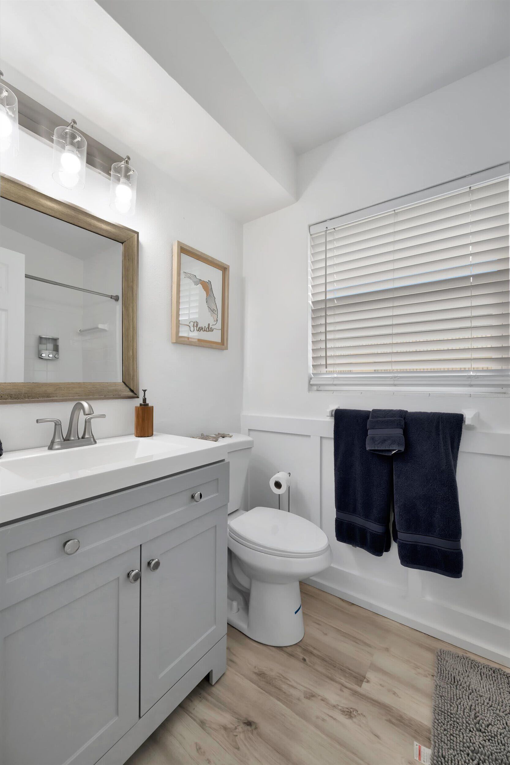A bathroom with a white vanity, a large mirror, a toilet, and a window with blinds.