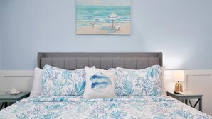 Um quarto com cama, mesa de cabeceira, abajur e pintura de uma praia.