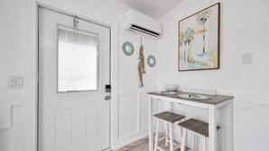 Uma porta branca com janela, um ar condicionado, uma pequena mesa com bancos e um quadro na parede.