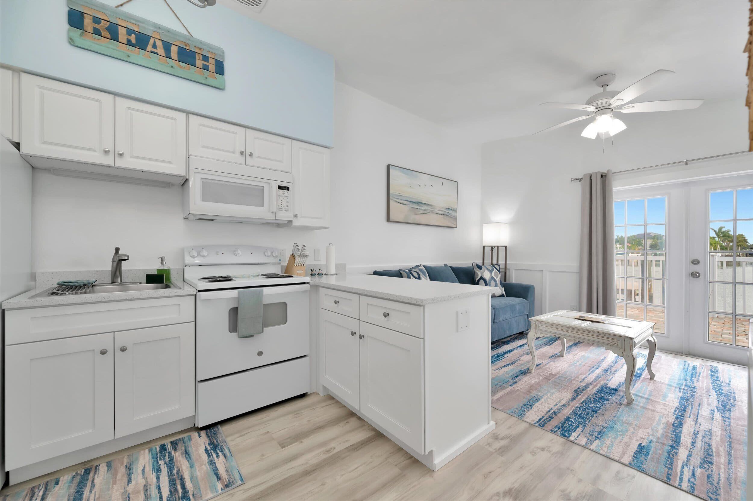 A modern kitchen with white cabinets, a microwave, and an oven. There's a blue couch and a small table in the living area.