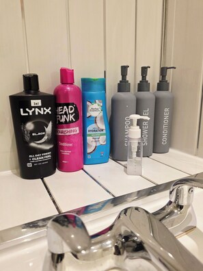 Une étagère de salle de bain contenant divers produits de toilette, notamment un flacon noir étiqueté « LYNX », un flacon rose étiqueté « HEAD FUNK », un flacon bleu étiqueté « Herbal Essences » et deux flacons gris étiquetés « GEL DOUCHE » et « APRÈS-SHAMPOING ».