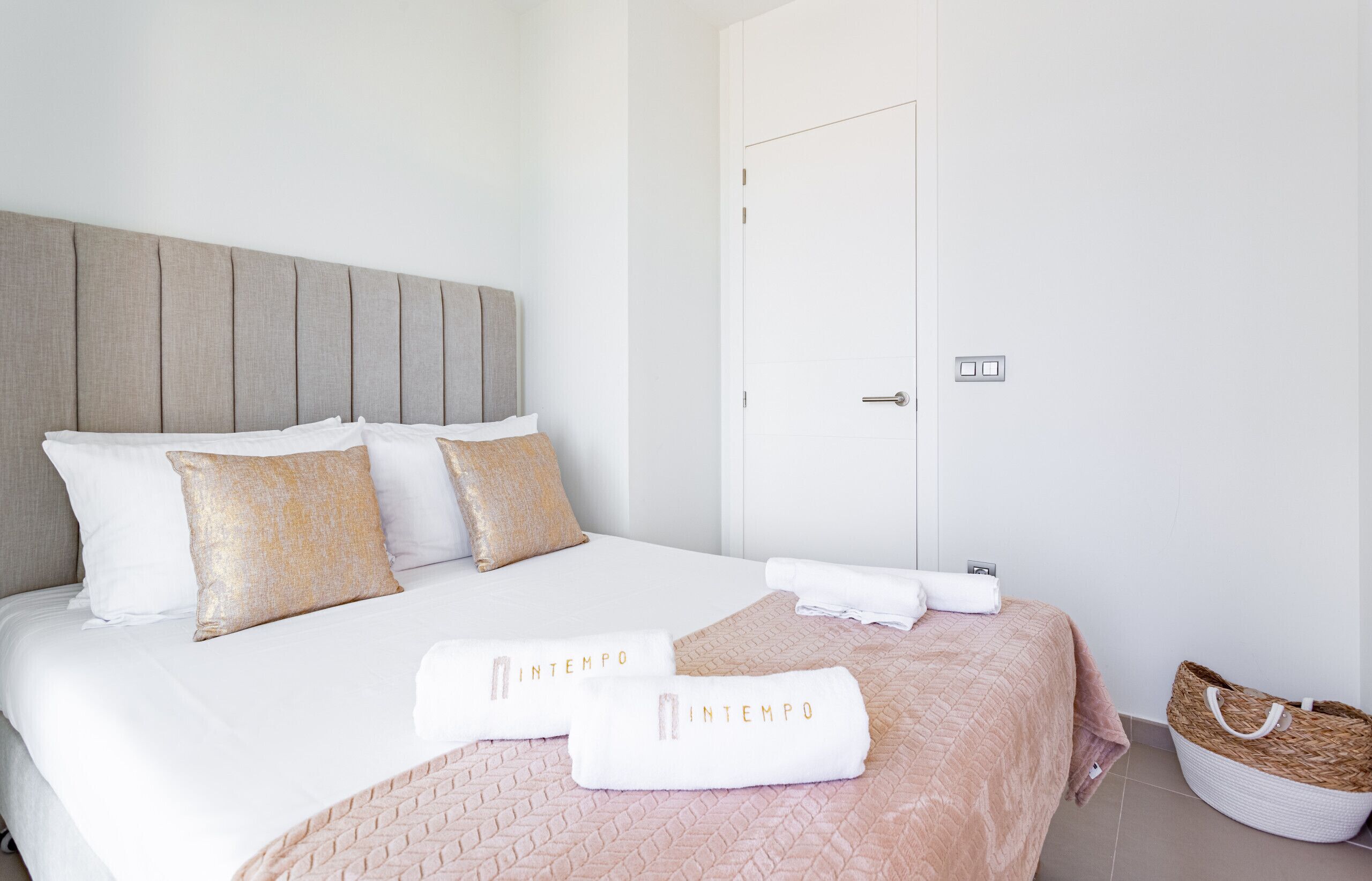 A neatly made bed with white and beige bedding, two towels with 'INTEMPO' text, and a woven basket on the floor.