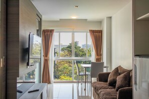 A modern living room with a flat-screen TV, a sofa, a dining table with chairs, and a large window with curtains. - DWIJA HOTEL & SUITE (Pattaya)