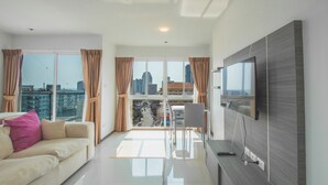 Una sala de estar moderna con un sofá, un televisor y vistas de la ciudad a través de ventanas amplias.