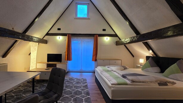 Une chambre confortable située dans les combles, avec un lit, un bureau et une chaise, des poutres apparentes et une petite fenêtre.