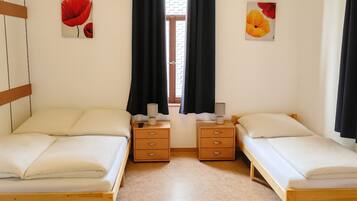 Quarto duplo com duas camas, cada uma com estrutura de madeira e roupa de cama branca. Possui duas mesas de cabeceira com abajures e uma pequena gaveta. O quarto tem uma janela com cortinas pretas e vista para um muro de tijolos lá fora.