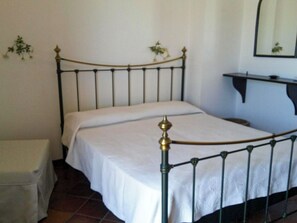 Un letto singolo con struttura in metallo e copriletto bianco in una stanza con pareti bianche e pavimento piastrellato.