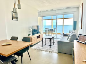 Ein modernes Wohnzimmer mit Essbereich, Fernseher und Meerblick.