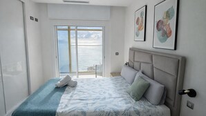 Une chambre à coucher avec un lit, une vue sur la mer par une porte coulissante et deux œuvres d’art encadrées au mur.