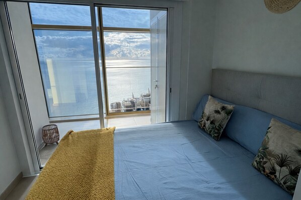Et soveværelse med en stor skydedør, der fører ud til en balkon med udsigt over havet og bygninger.