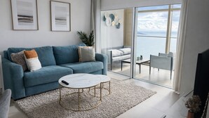 Un salon moderne avec un canapé bleu, une table basse blanche et une vue sur la mer, visible à travers de grandes portes vitrées.