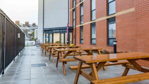 An outdoor area with multiple wooden picnic tables arranged in rows, a brick building with large windows, and a modern building with a glass facade in the background. - Student Only Mary Parker House (Plymouth)