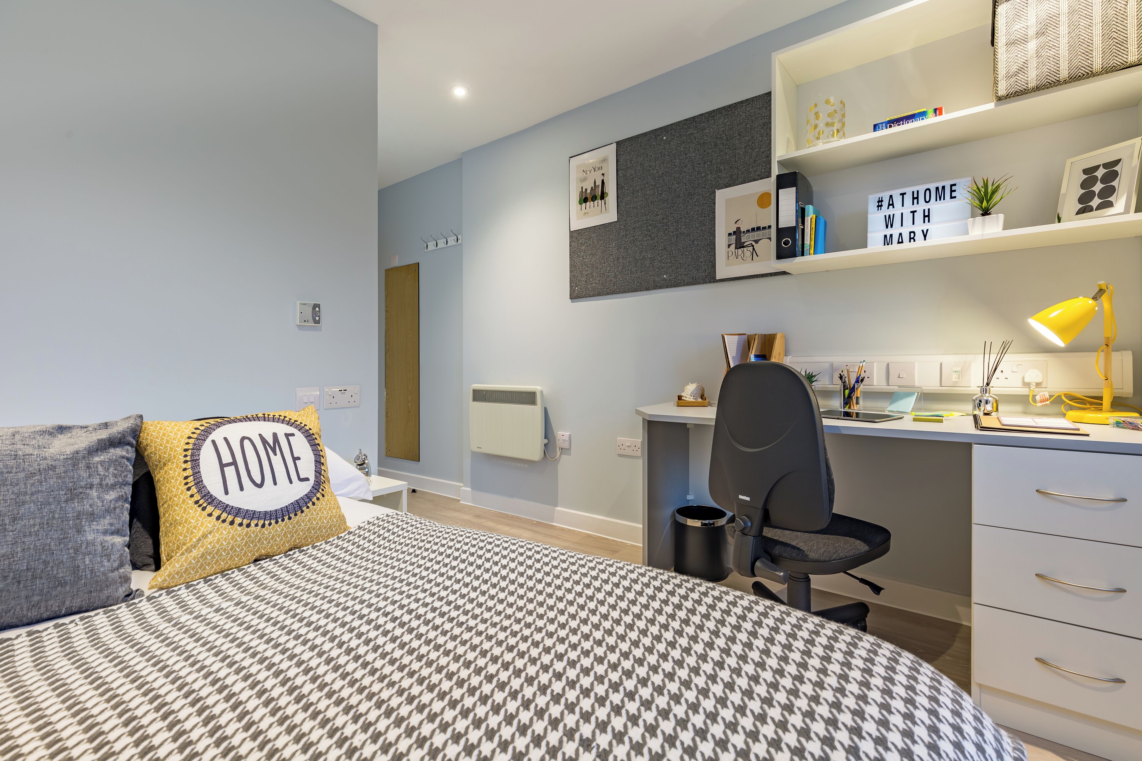 A bedroom with a bed, a desk, and a chair. There is a radiator, a shelf with books and decorative items, and a wall with framed pictures.