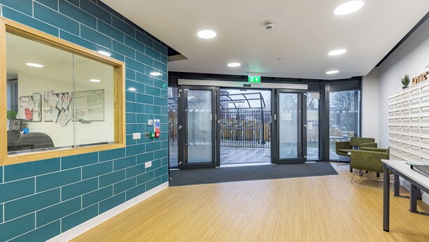 A reception area with a glass door entrance, teal tiled walls, a wooden floor, and a seating area with green chairs. - Student Only Mary Parker House (Plymouth)