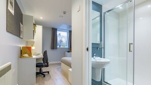 A modern bedroom with a desk, chair, and a bed. There is a bathroom with a glass shower and a sink. - Student Only Mary Parker House (Plymouth)