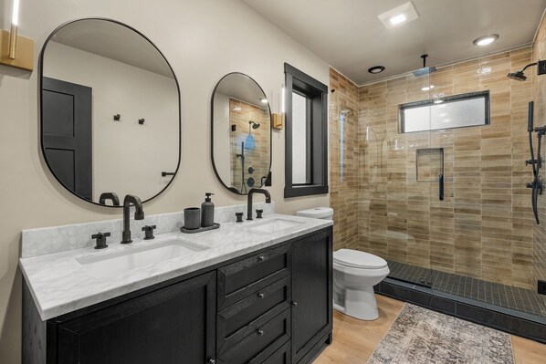 A modern bathroom with a large mirror, two sinks, a toilet, and a shower area. - Skycrest Heights ~ Indoor Pool ~ Firepit ~ Unbelievable Views! (Sevierville)