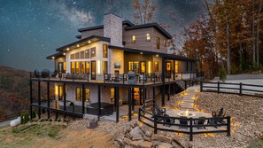 A modern two-story house with a stone facade, large windows, and a deck with outdoor seating. - Skycrest Heights ~ Indoor Pool ~ Firepit ~ Unbelievable Views! (Sevierville)