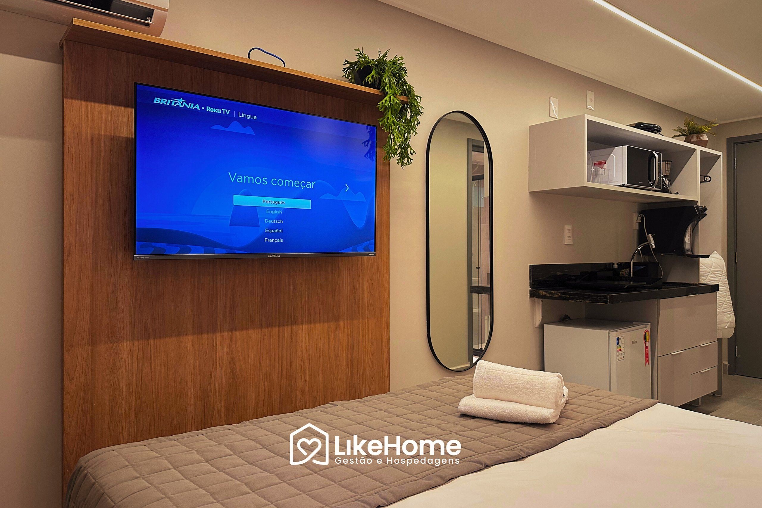A hotel room with a television displaying a language selection screen, a bed with a quilted cover, a bedside table with a towel, a mirror, and a kitchenette area.