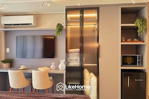 A modern living room with a flat-screen TV, a built-in air conditioner, a dining area with chairs, a sofa, and a kitchen area with a microwave and refrigerator.