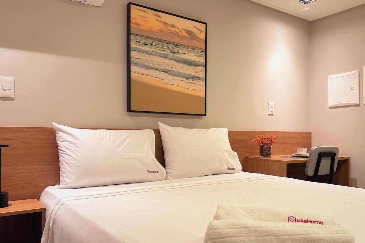 Ett hotellrum med en säng, två kuddar, ett nattduksbord, ett skrivbord, en stol, en tavla som föreställer en strandscen och en handduk med en logotyp.