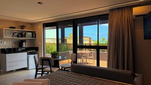 Habitación de hotel moderna con kitchenette, una cama grande, zona de comedor con sillas y un balcón con vistas a edificios y zonas verdes.