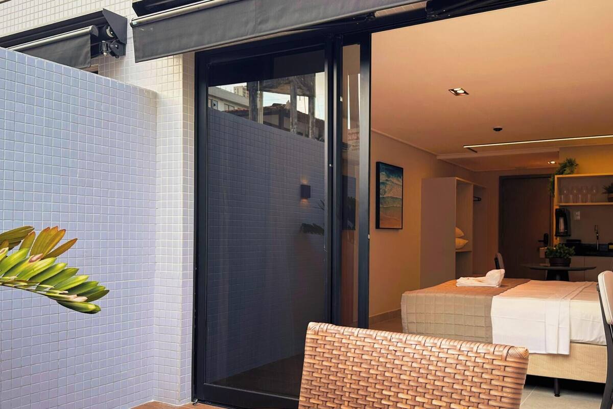 Ett modernt hotellrum med en skjutdörr i glas som leder ut till en balkong, en säng med ett vitt överkast, ett litet skrivbord med en stol och en liten köksdel.