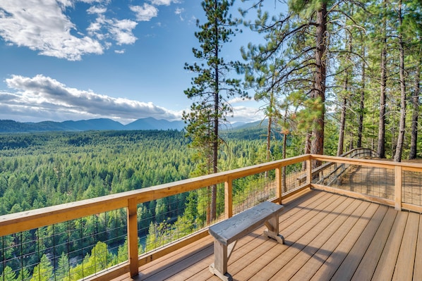 A wooden deck with a bench, overlooking a forested valley and mountains.