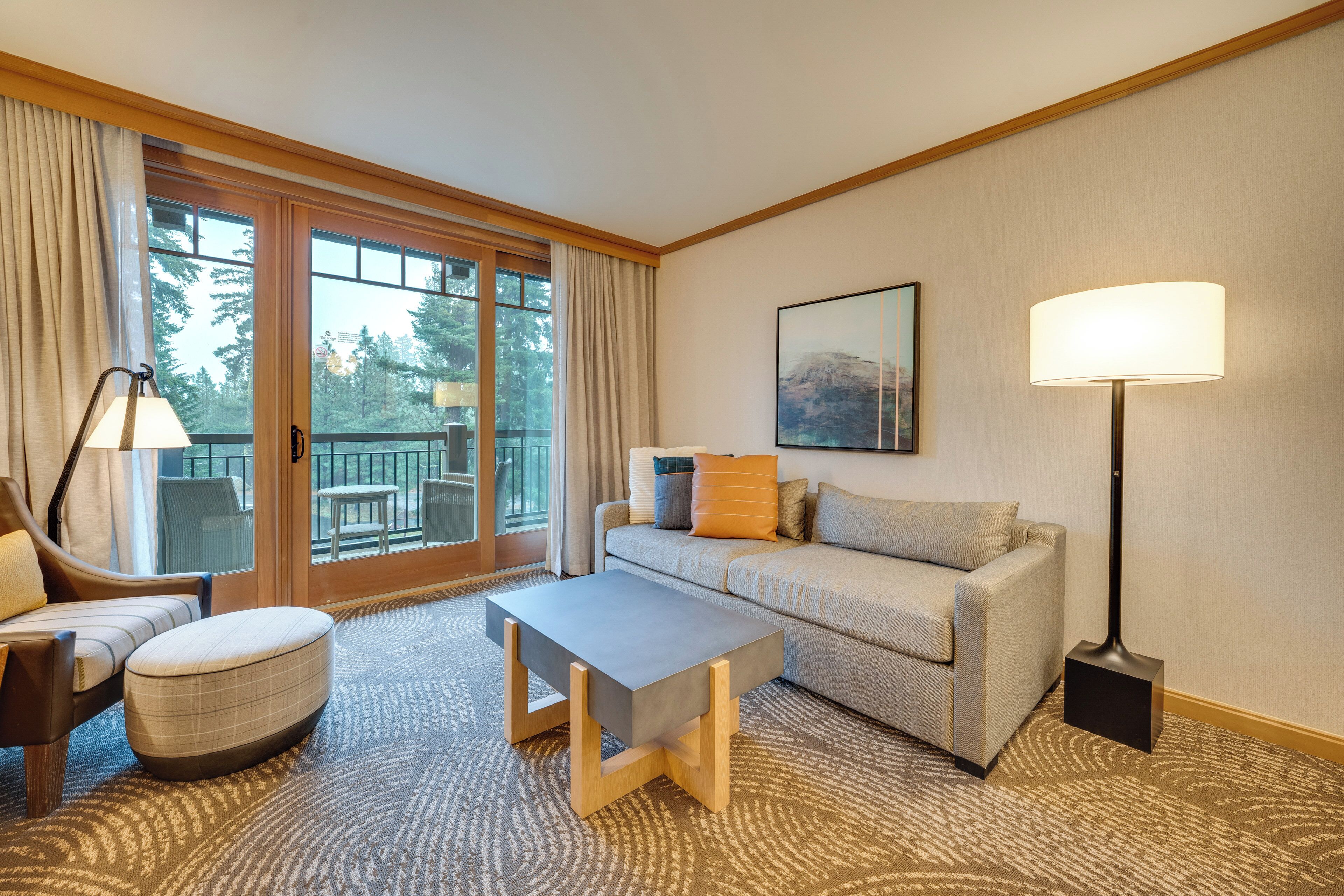 A modern hotel room with a sofa, armchair, ottoman, and a small table. There is a large window with curtains, a floor lamp, and a painting on the wall.