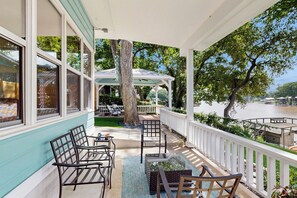 A covered outdoor seating area with chairs and tables, overlooking a body of water and trees. - Dock, Patio & Grill! Waterfront Lake Dunlap Home (New Braunfels)