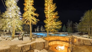 A stone fire pit surrounded by snow, with trees adorned with lights and a pool in the background. - Pool, Hot Tub & Golf: Everline Resort Corner Suite (Olympic Valley)