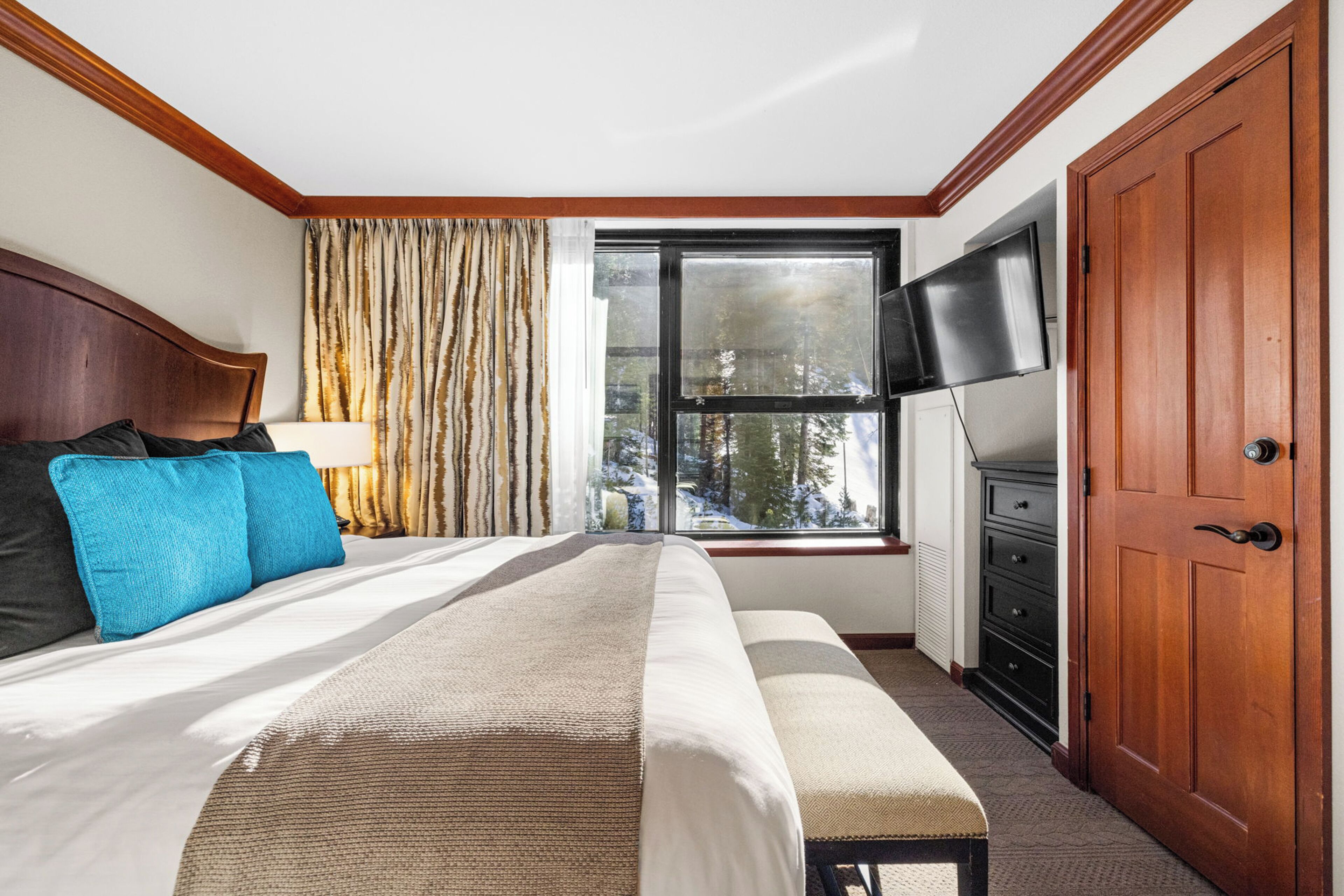 Habitación de hotel con una cama, un banco, un televisor, un ventanal con vistas a árboles y una puerta de madera.