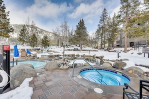 Zona de piscina con dos jacuzzis rodeados de rocas y nieve, un flotador salvavidas y un cartel que indica un interruptor de emergencia.