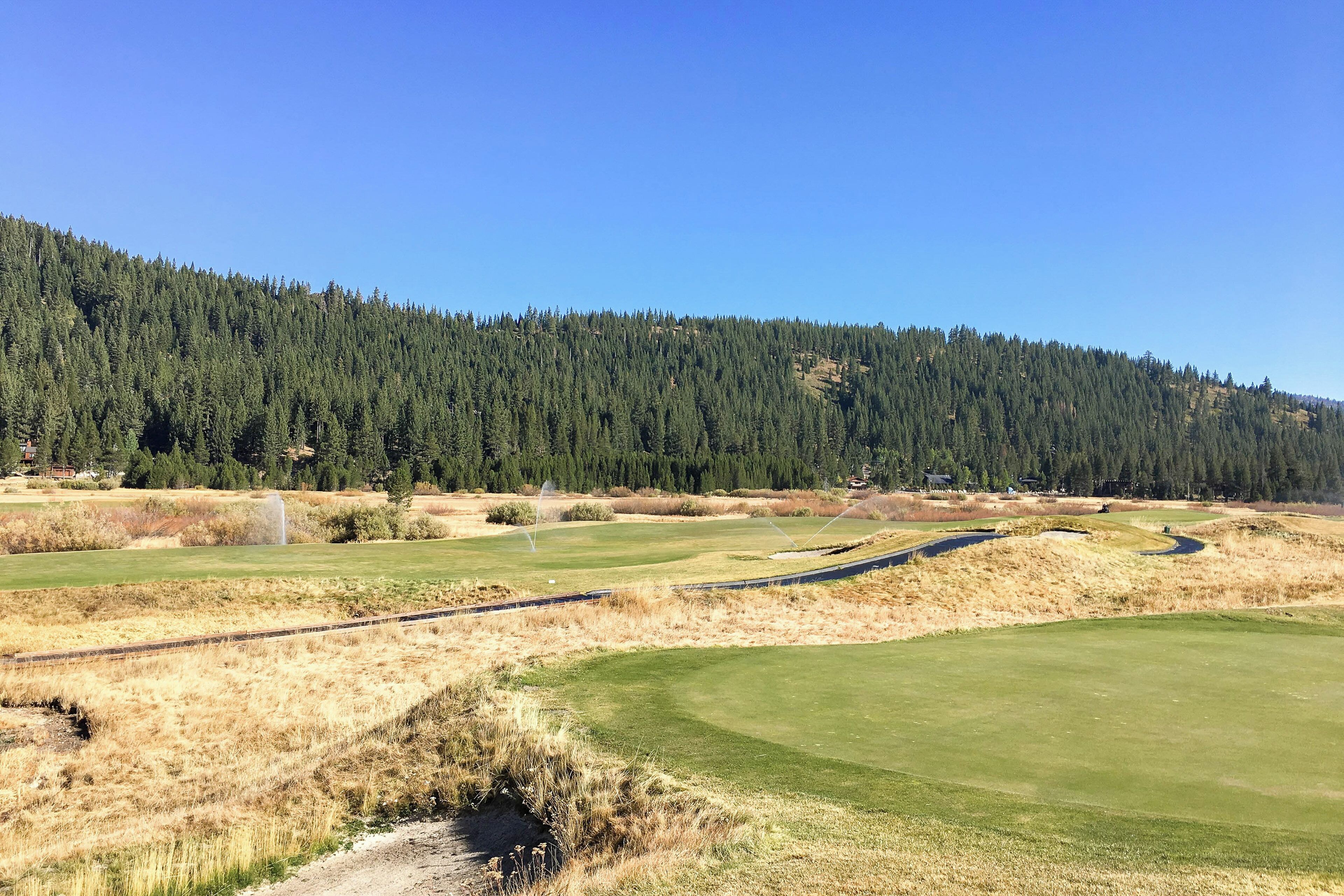 A golf course with a clear path, a water feature, and a forested hillside in the background.