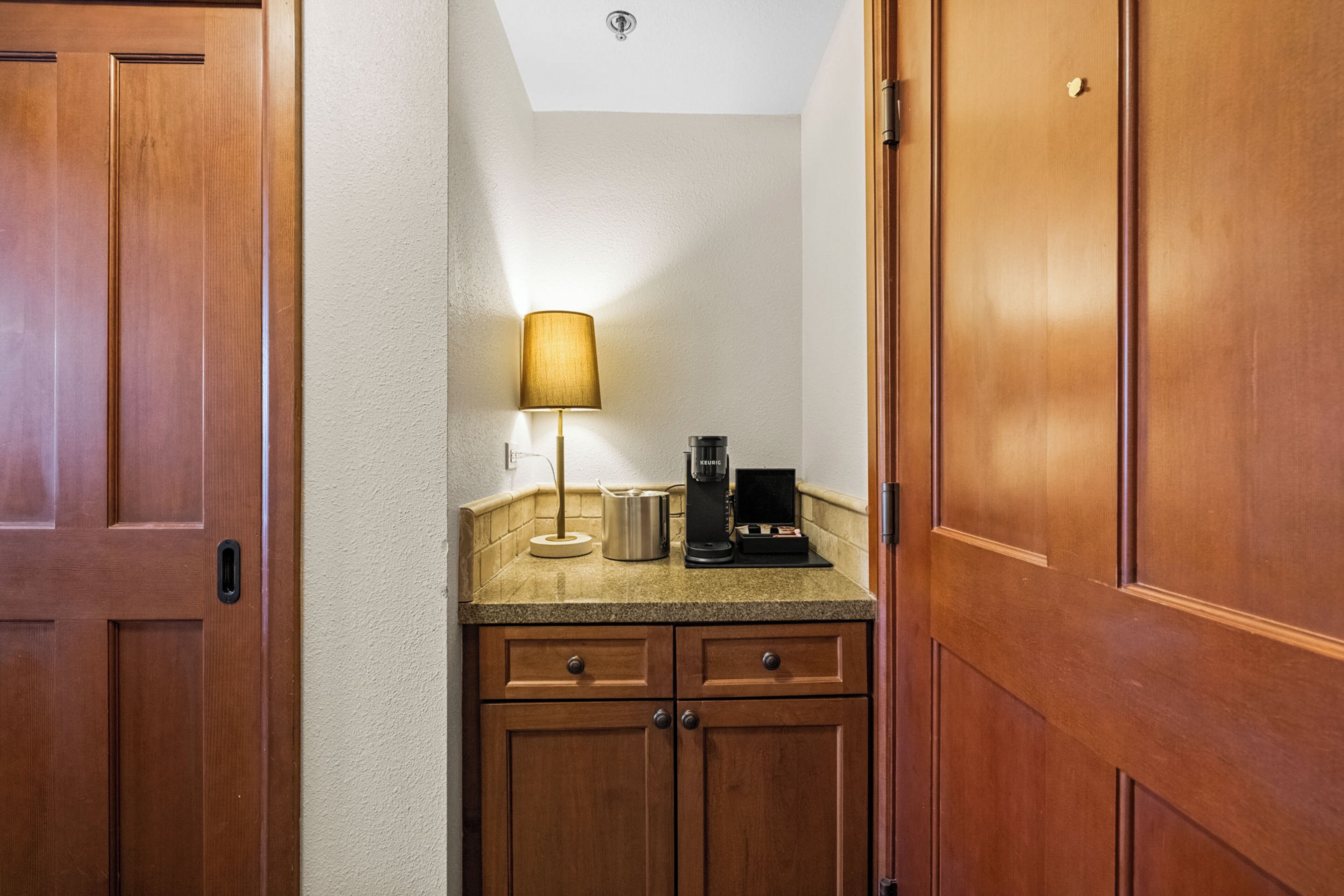A small kitchenette with wooden cabinets, a coffee maker, and a lamp on the countertop.
