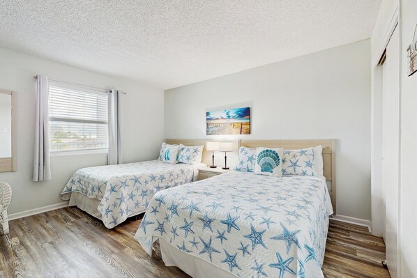A bedroom with two beds, a window with curtains, a painting on the wall, and a wooden floor. - 2 BR Condo w/ Shared Pool & Beach Access (Fort Walton Beach)