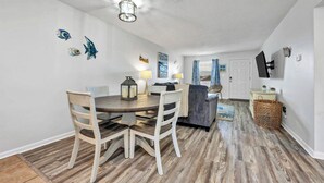 A dining area with a wooden table and chairs, a lamp, and wall art. - 2 BR Condo w/ Shared Pool & Beach Access (Fort Walton Beach)