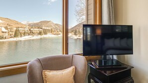 Ein Zimmer mit großem Fenster mit Blick auf einen See und Berge, einem Standfernseher und einem gepolsterten Sessel.