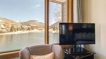 A room with a large window overlooking a lake and mountains, a flat-screen TV on a stand, and a cushioned chair.