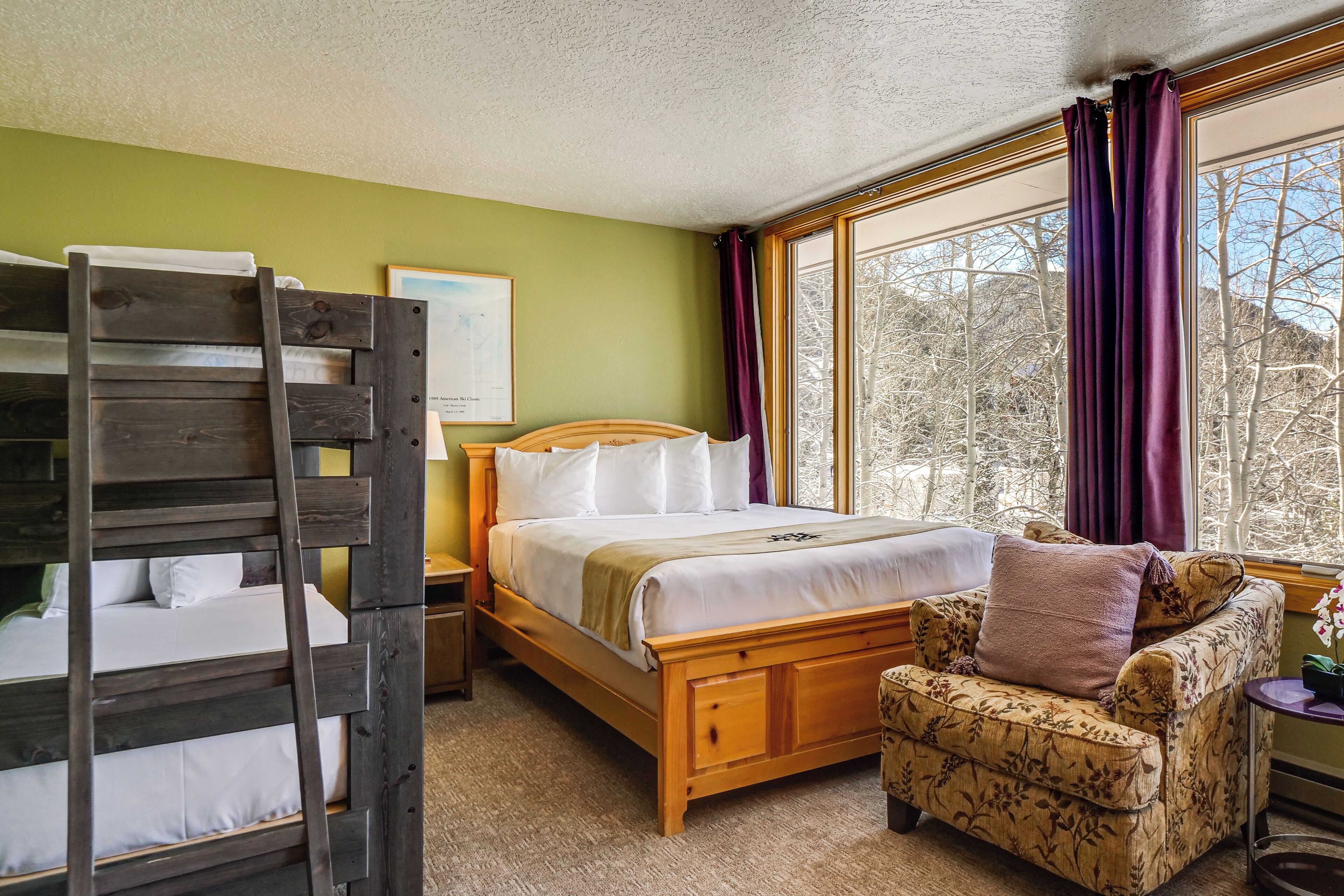 A bedroom with a wooden bunk bed, a single bed with white bedding, a floral armchair, a nightstand, and a window with purple curtains.