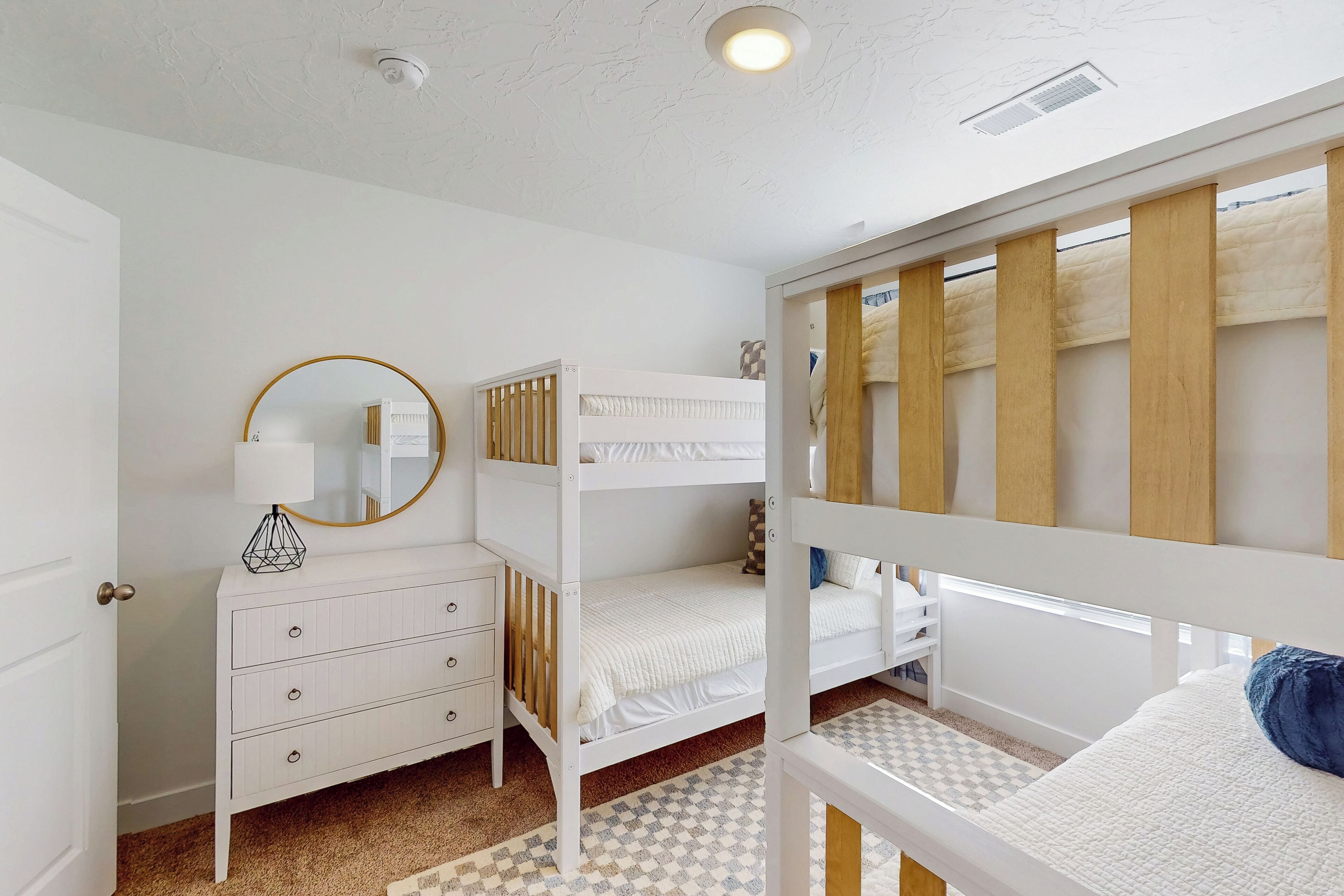 Une chambre avec des lits superposés, une commode blanche, un miroir et un tapis à motifs.