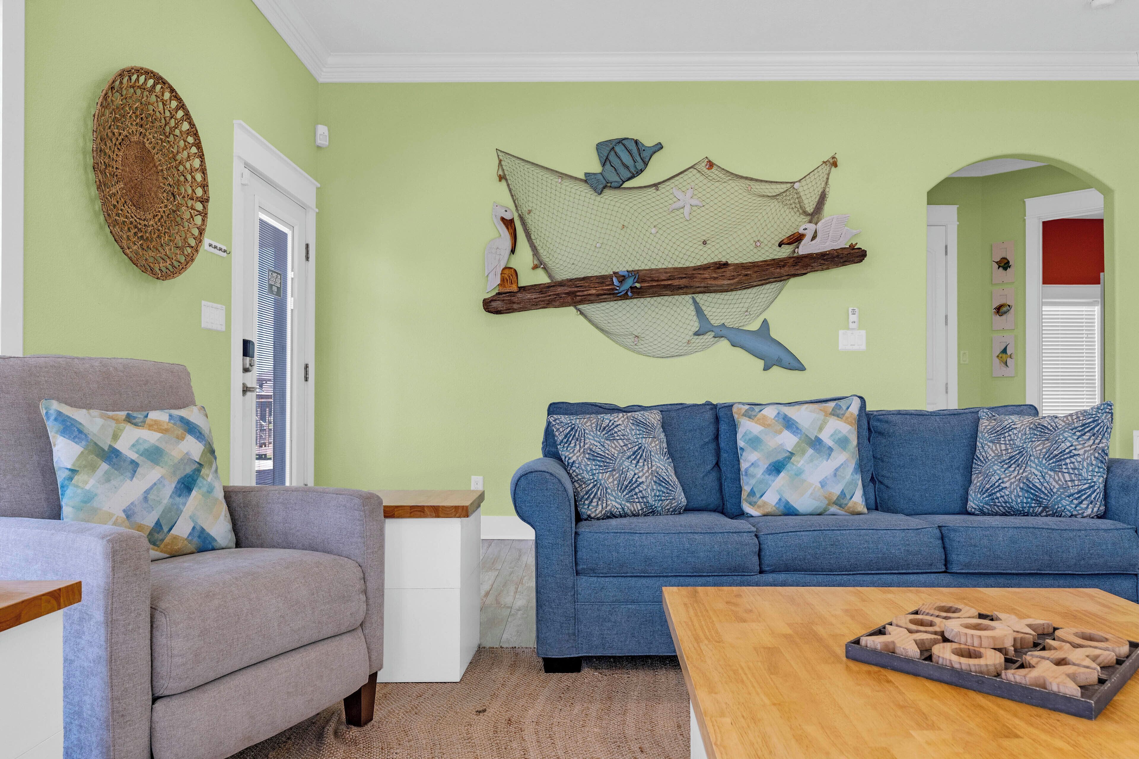 A living room with a blue sofa, a grey armchair, a wooden coffee table, and a wall hanging with a birdcage and fish.
